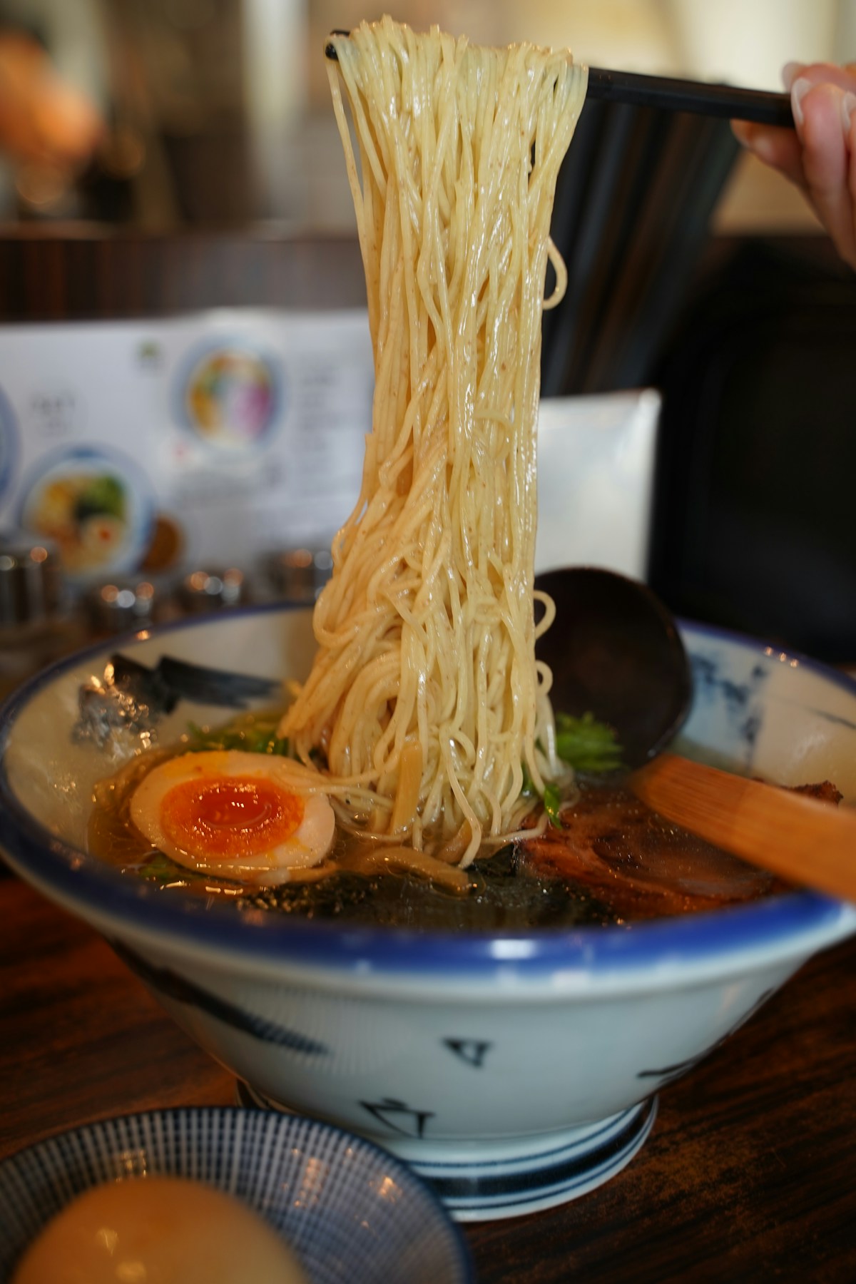 Thin noodles lifted high out of a bowl of ramen with a soft egg