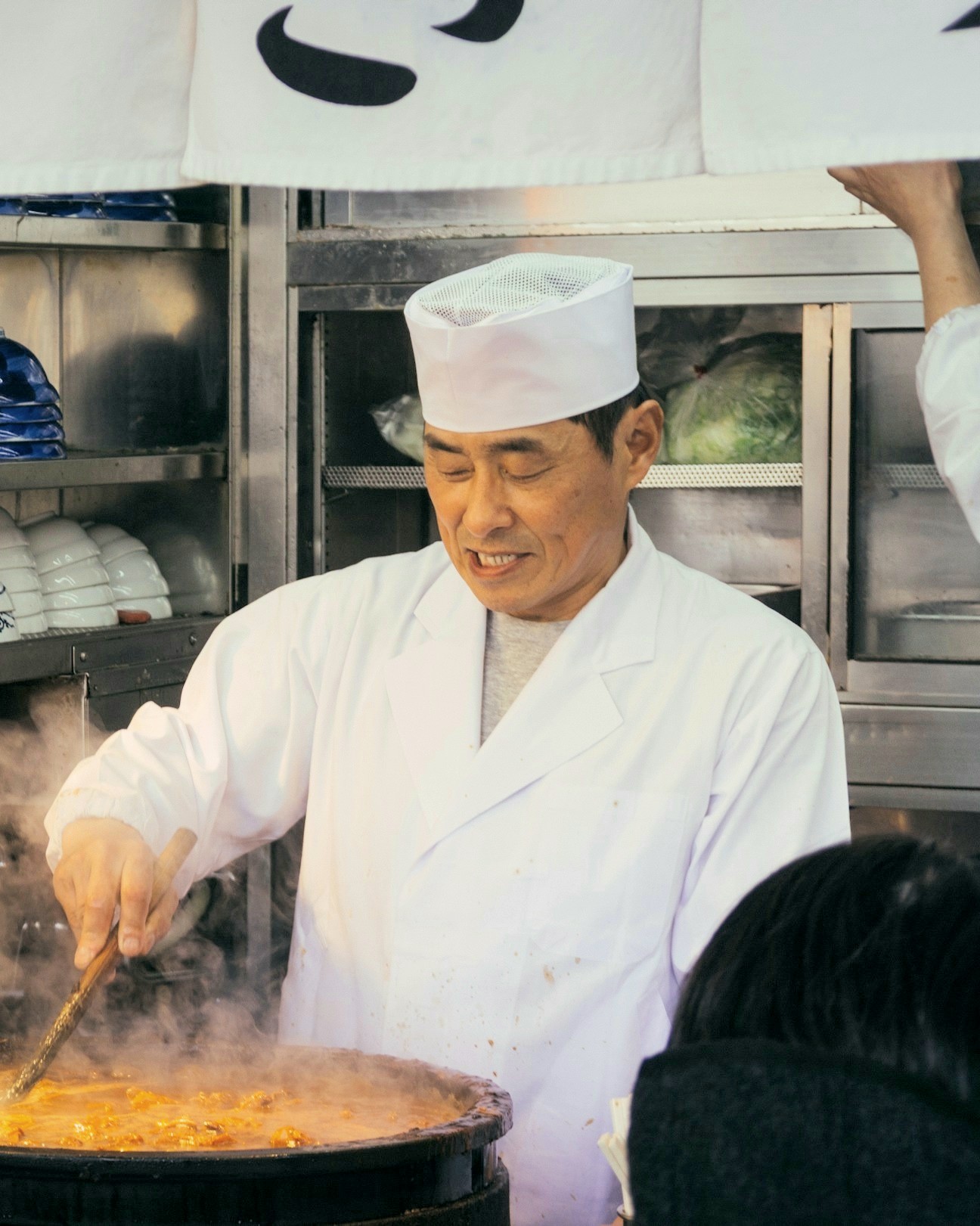 A cook in whites stirs a steaming pot at the ramen counter while guests eat
