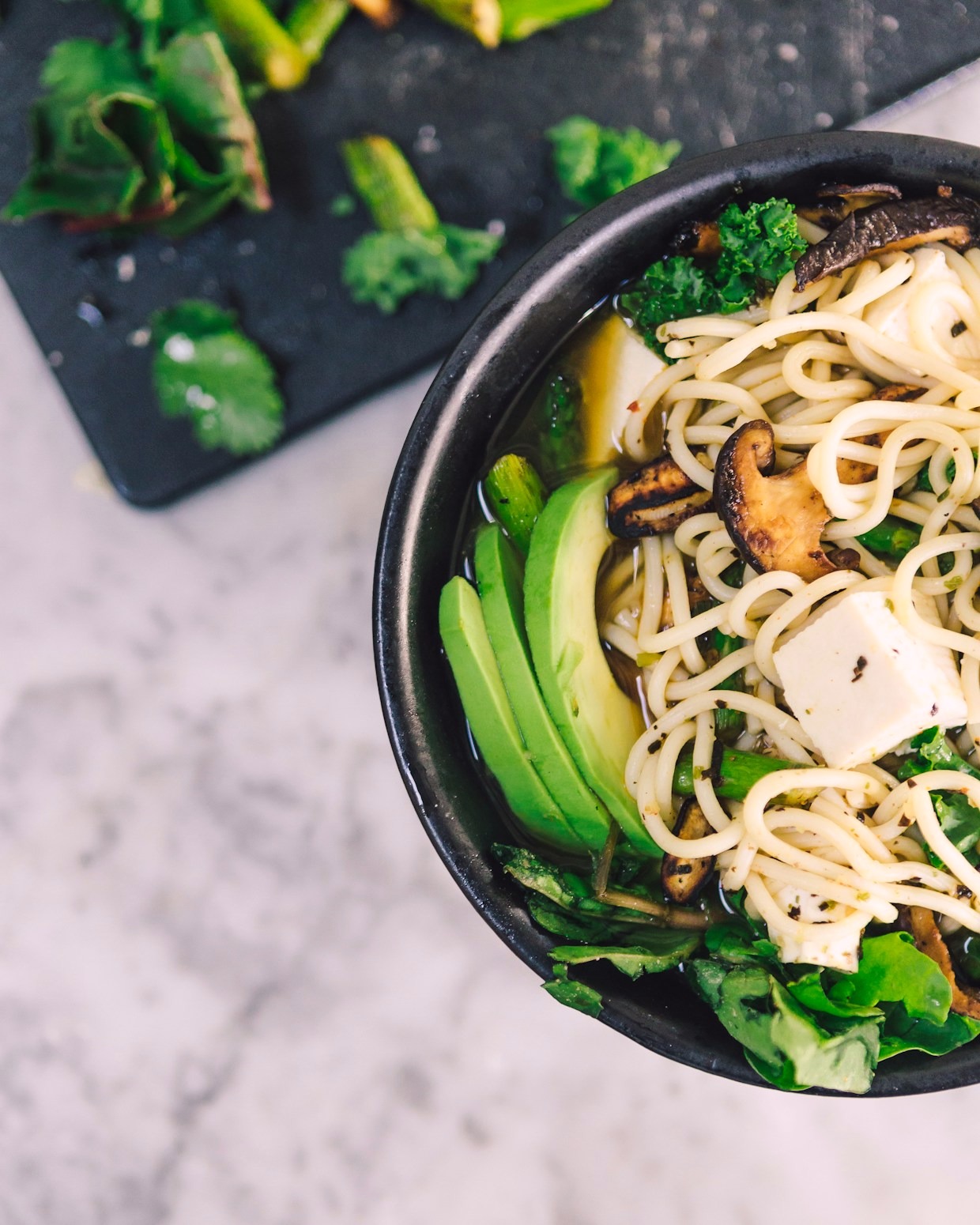 A vegan miso ramen with silken tofu, shiitake, avocado and greens in a black bowl