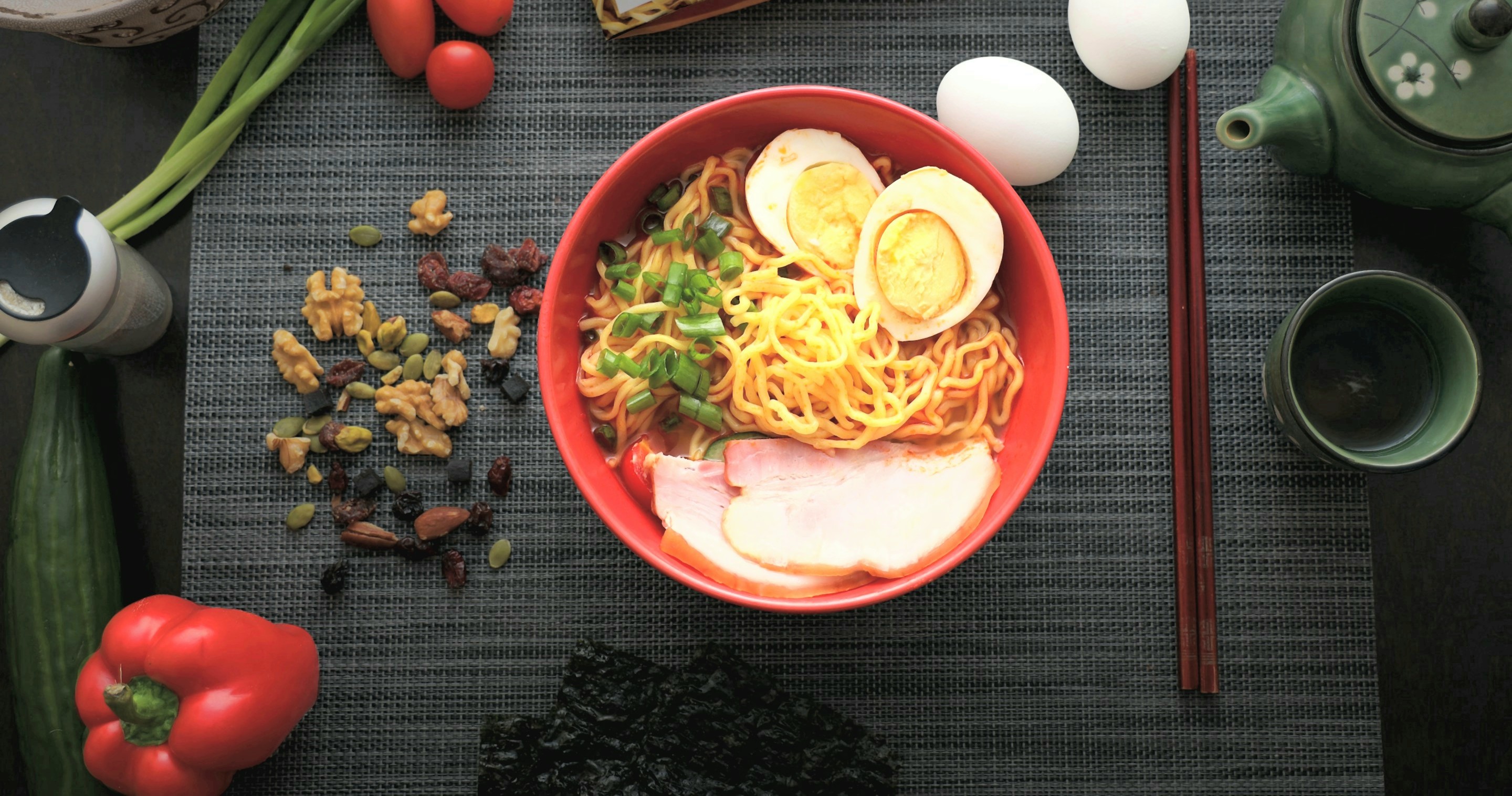 A red bowl of ramen with soft eggs, chashu and scallions on a dark mat, with chopsticks, a teapot and fresh vegetables around it