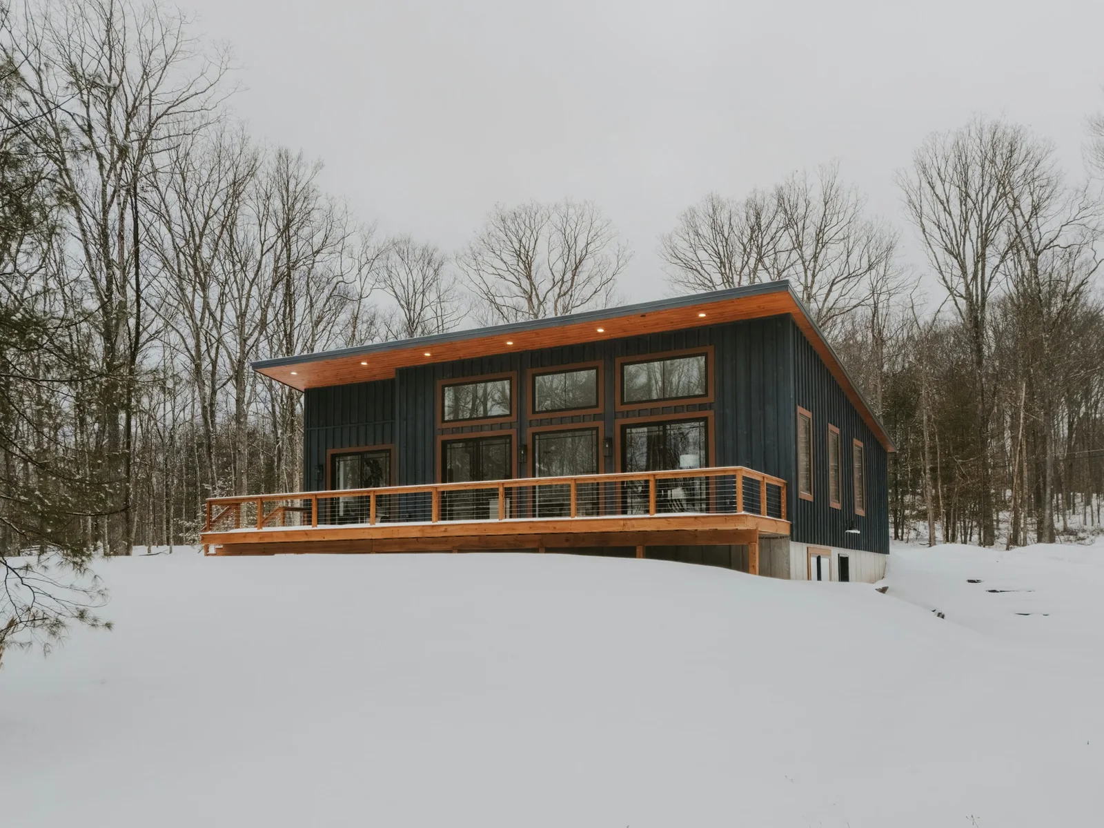 Fell House: a dark timber house with a long deck, raised above a snowy hillside
