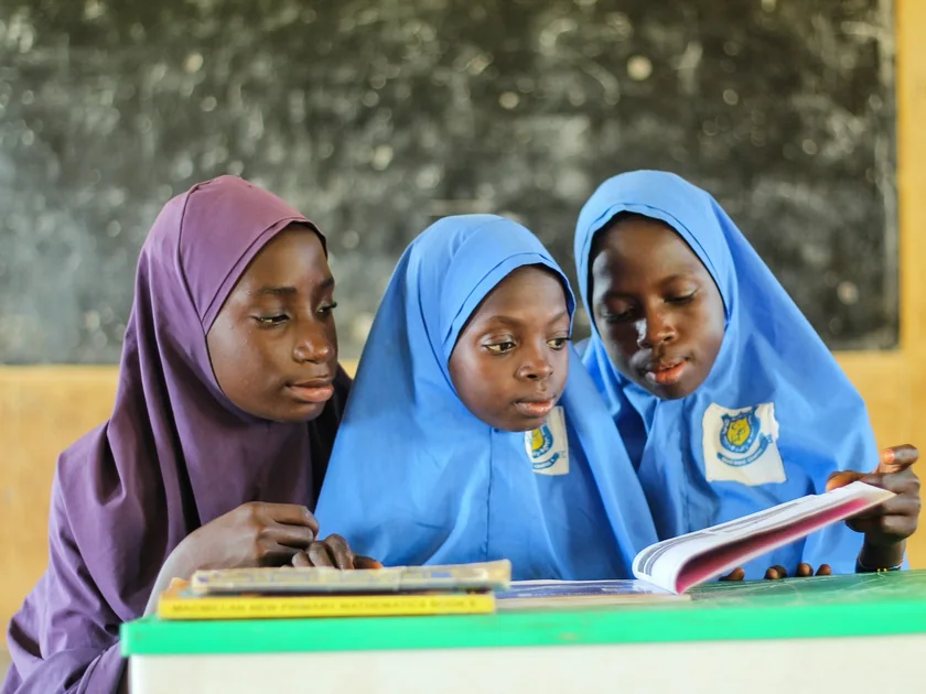 Three girls in school hijabs read a textbook together at their desk