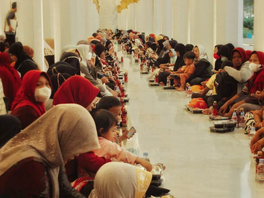 Families sit in long rows on the floor of a bright hall, waiting to open their fast