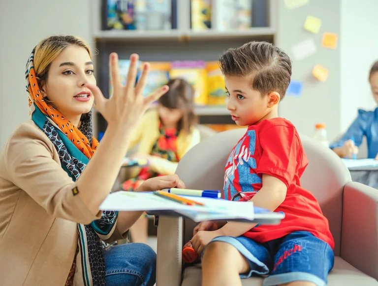 A teacher explains a lesson to a young boy at his desk in a bright classroom