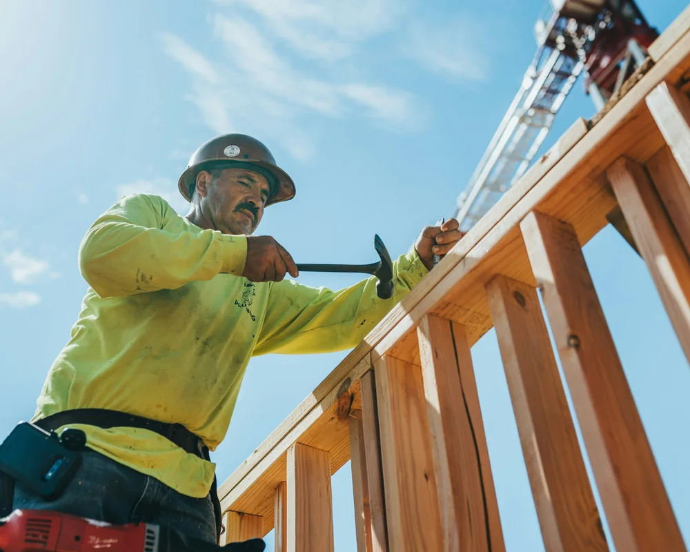 A carpenter in a hard hat nailing a timber frame against a blue sky