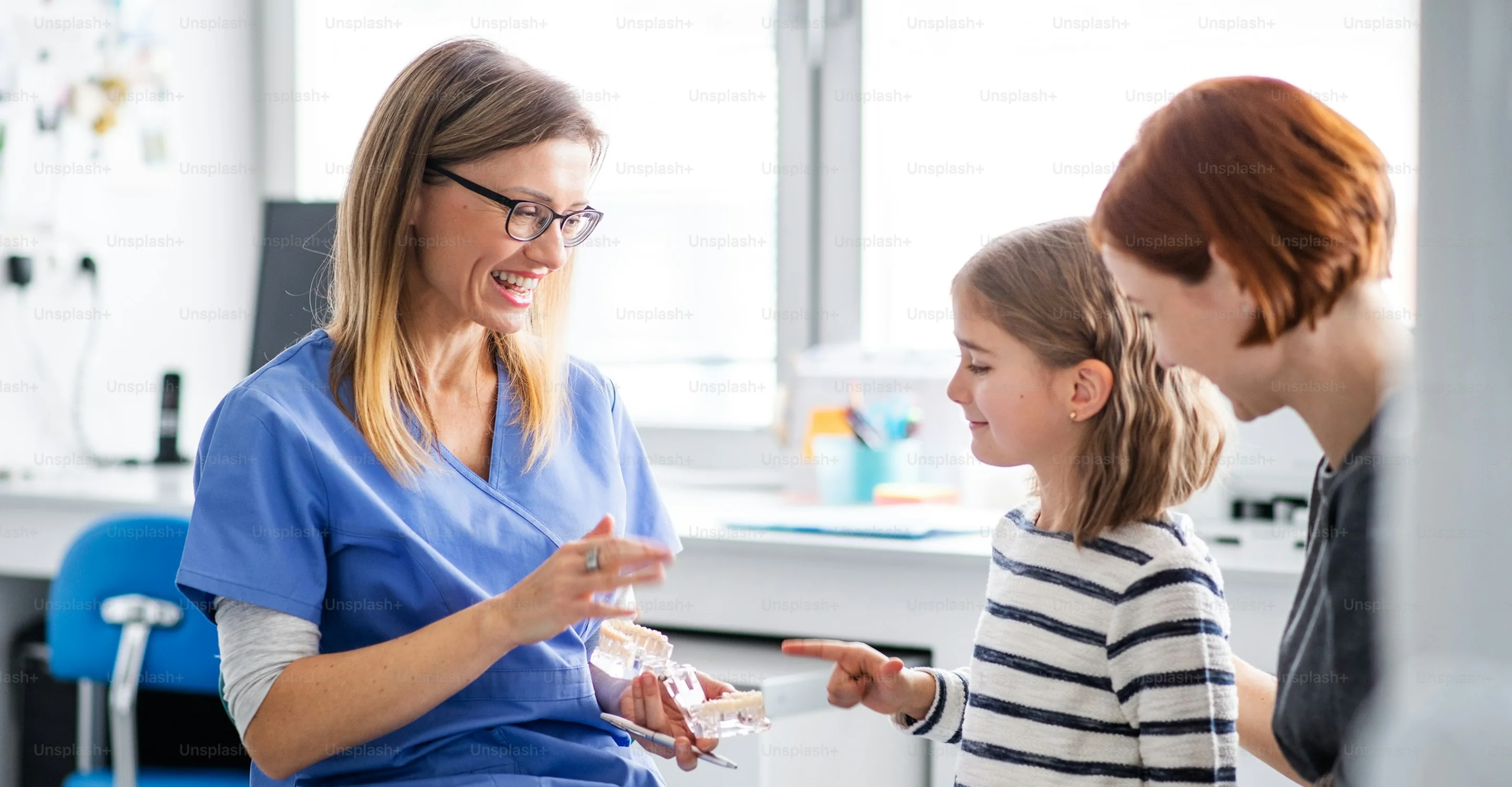 A smiling dentist shows a girl a model of teeth while her mom watches in a bright clinic room