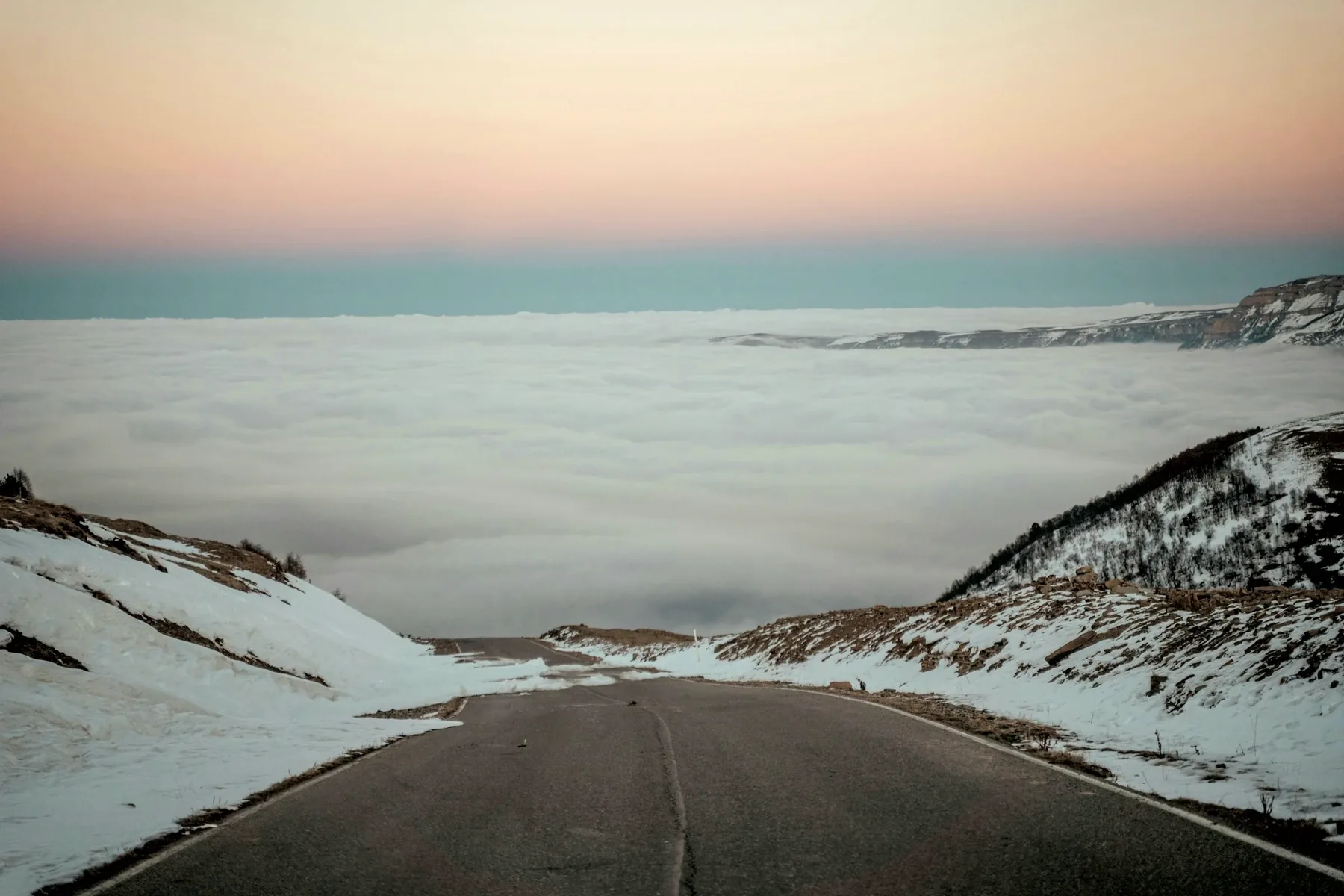 A cleared mountain road between snowbanks running down into a sea of fog at dawn