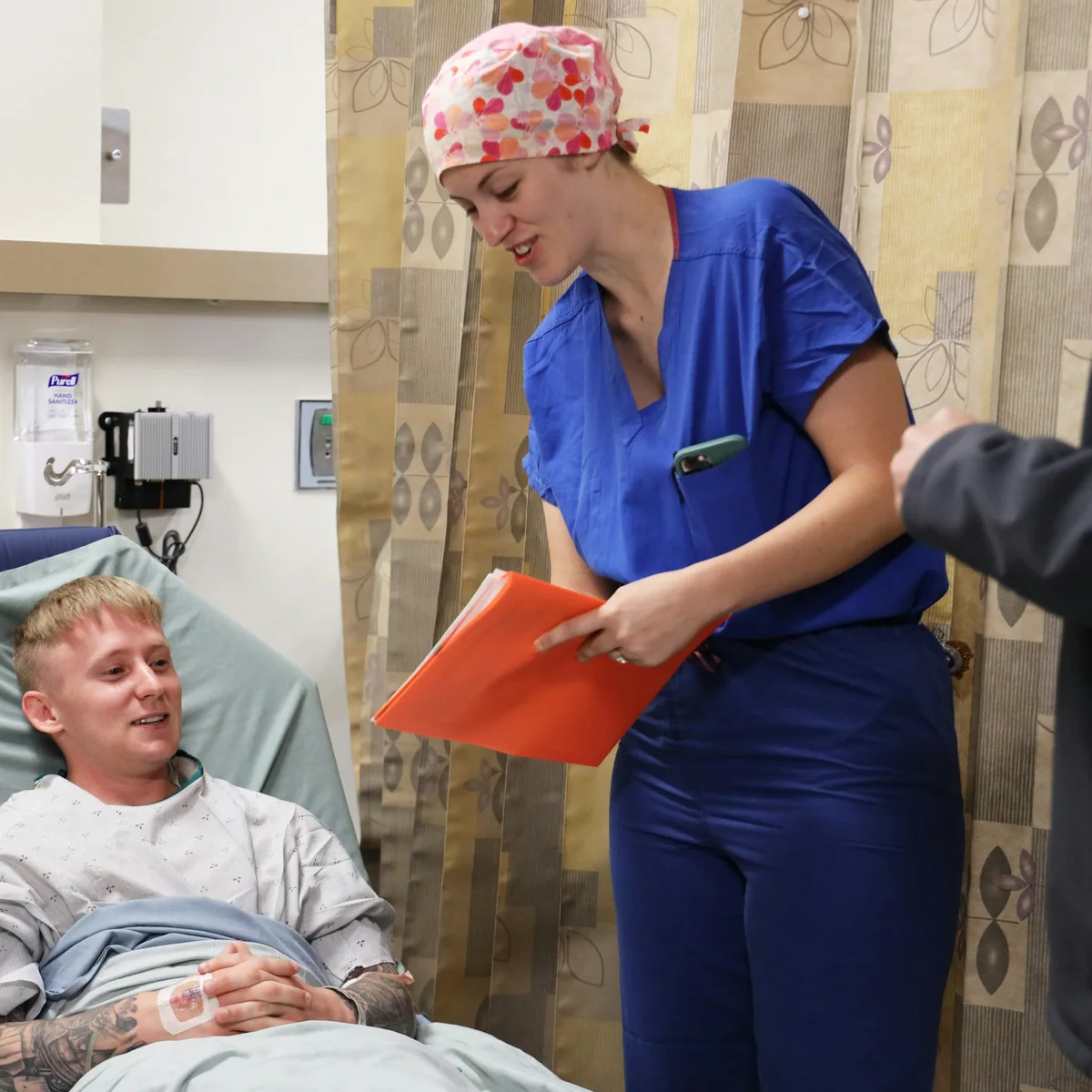 Nurse in blue scrubs checking notes beside a smiling patient on a ward